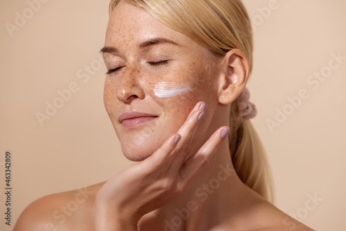 Close up of beautiful young female with cream on a face. Smiling woman with closed eyes touching cheek with fingers.