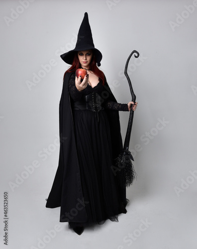 Full length portrait of dark haired woman wearing black victorian witch costume. standing pose, holding a broomstick with gestural hand movements, against studio background.