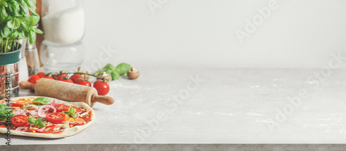 Homemade pizza background with wooden rolling pin, flour and basil leaves on grey kitchen table at white wall background. Cooking Italian food at home. Front view with copy space.
