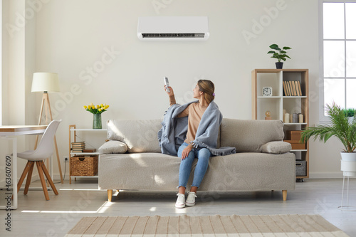 Woman who's sitting on sofa under warm plaid in living room switches off her air conditioner on wall. Young girl adjusting modern AC system, regulating temperature and enjoying cool fresh air at home