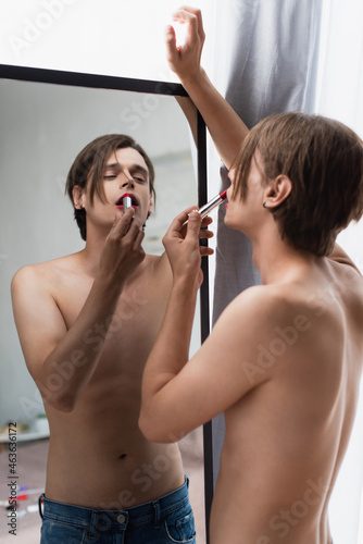 shirtless transgender young man applying red lipstick and looking at mirror at home