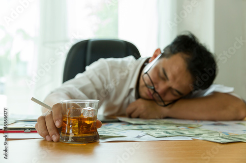 Stressed adult Asian businessman at workplace and drinking alcohol with many document paper for business on desk. Asian man overstressed at job while sitting at office desk in the office