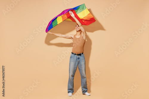 Full body young activist queer happy fun blond latin gay man with make up in beige tank shirt waving hold rainbow flag isolated on plain light ocher background studio People lgbt lifestyle concept