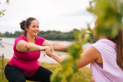 Medium shot of professional fitness female trainer giving personal training to overweight young woman outdoor in summer day. Instructor help fat woman lose weight outside doing squats outside.