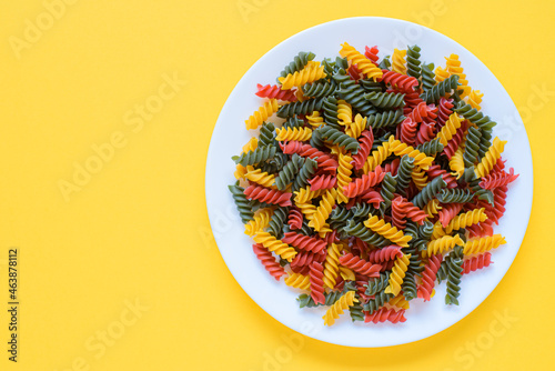 Red green and yellow dry raw fusilli pasta on a plate with yellow background and copy space.