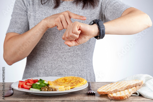Intermittent fasting concept with a woman sitting hungry in front of food and looking at her watch to make sure she breaks fast on the correct time. A dietary modification for healthy lifestyle.