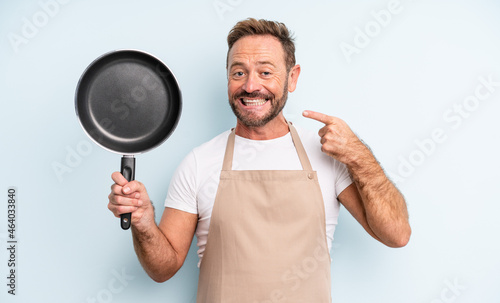 middle age handsome man smiling confidently pointing to own broad smile. frying pan concept