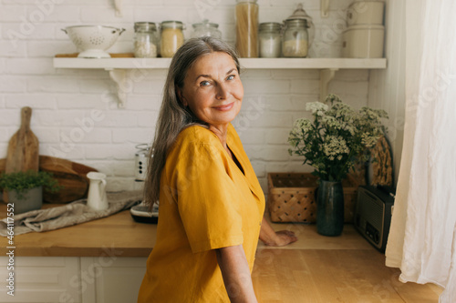 Lovely senior woman in yellow dress posing in countryhouse. Smiling positive female going to cook on kitchen, look from window. Weekends away, nature, interior, pure joy, growing old concept