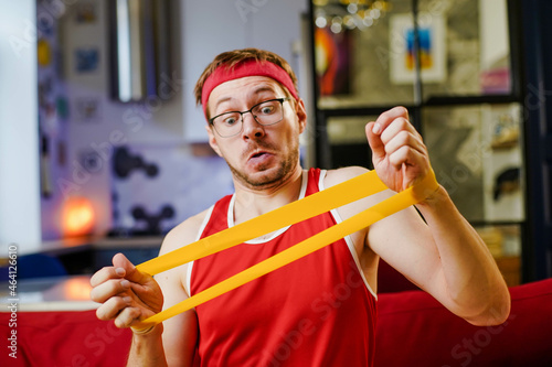 Portrait of funny man in red retro sportswear doing exercises with an elastic resistance band at home.