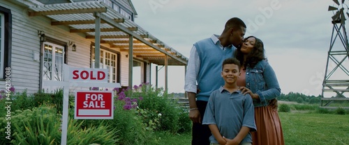 Portrait of happy African American Black family posing near sold sign, their new house in the background