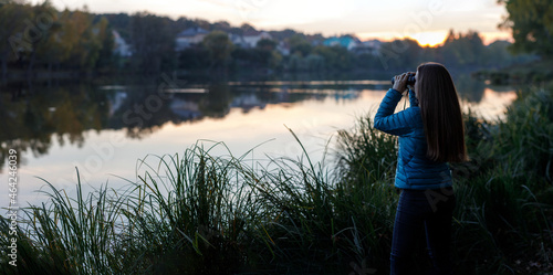 Young birdwatcher woman standing with binoculars near the pond in the evening