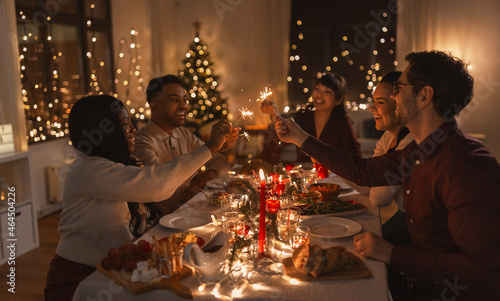 holidays and celebration concept - multiethnic group of happy friends with sparklers having christmas dinner at home