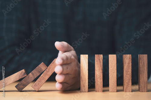 ฺBusinessman uses his hand to stop a falling wooden block to a standing wooden block domino , It is a symbol of protection against damage or stop loss for crisis management concept.