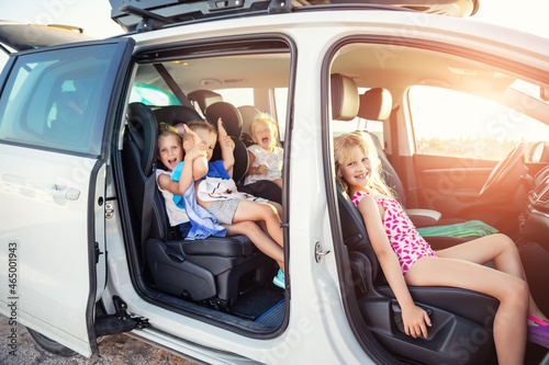 Group of four cute adorable little happy caucasian children enjoy having fun sit in minivan going to sea beach road trip on hot summer day. Kids in swimsuit in car with side open door against sun