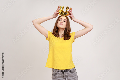 Portrait of selfish brown haired teenage girl in yellow T-shirt holding gold crown over head, having arrogance expression, privileged status. Indoor studio shot isolated on gray background.