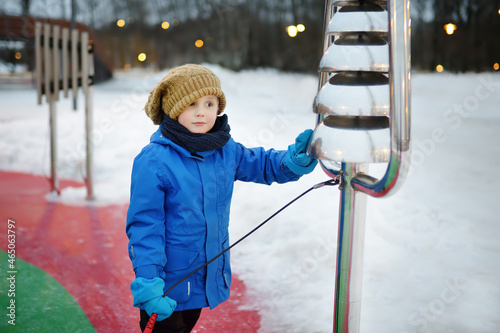 Little boy knocking by street xylophone drums on playground in public park on a snow winter day. Modern equipment of street city playgrounds.
