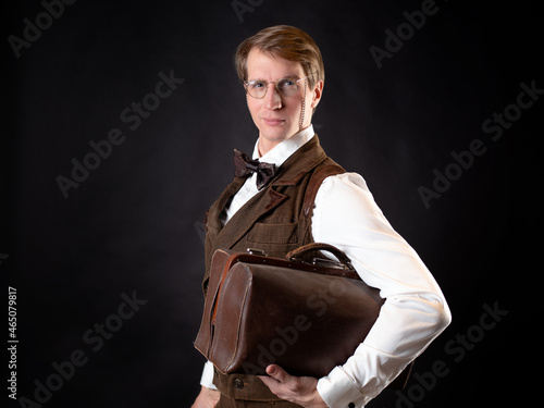 An intelligent gentleman in the Victorian style. Vintage retro suit, young attractive man in a vest and bow tie with glasses on a chain. he holds his bag in his hands