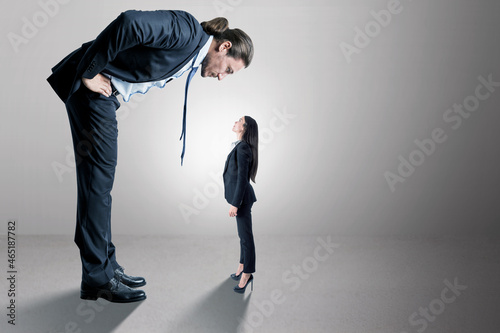 Businessman looking down on female subordinate on concrete background with mock up place for your advertisement. Boss and discrimination concept.