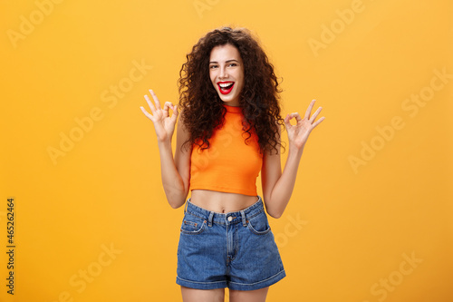 Delighted outgoing and friendly caucasian girl with curly hairstyle in cropped top and shorts showing okay gesture in approval expressing she like awesome party smiling at camera over orange wall