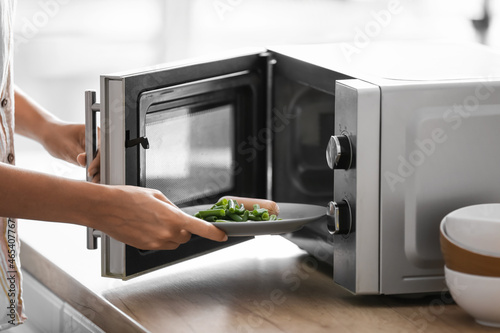 Young woman putting plate with food into microwave oven in kitchen, closeup