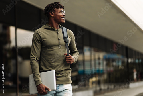 Handsome smiling dark-skinned student, wearing glasses with a backpack, holding a laptop, walking around the campus