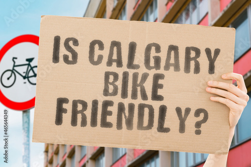 The question " Is Calgary bike friendly? " on a banner in men's hand with blurred background. Transportation. Zero waste. Bicycle lane. Streets. City. Safety. Insecure. Road signs. Dangerous