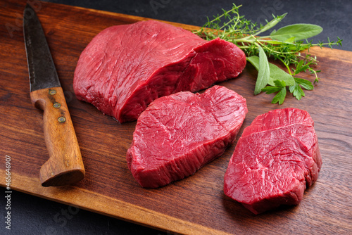 Raw dry aged bison beef rump steak piece and slices offered with herbs as close-up on wooden design board