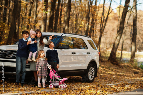 Mother stand near white suv car with four kids in autumn park. Family walk in fall forest.