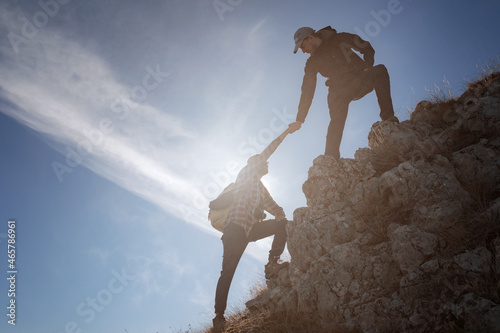 Silhouettes of two people climbing mountains and helping against the blue sky.