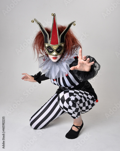 Full length portrait of red haired girl wearing a black and white clown jester costume, theatrical circus character. Sitting down on floor, isolated on studio background.