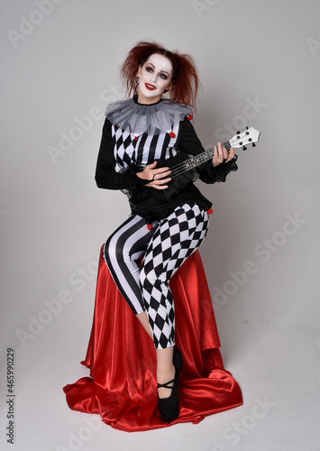 Full length portrait of red haired girl wearing a black and white clown jester costume, theatrical circus character. Sitting down on chair, isolated on studio background.