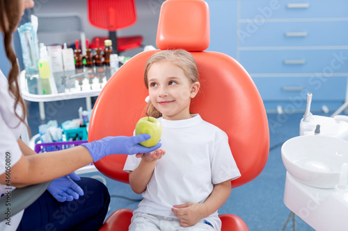 pediatric dentist gives a little girl a big green apple. beautiful girl is smiling in dentist's office. concept is a children's medical examination.