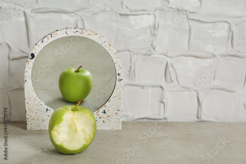 Bitten green apple near mirror with reflection of whole fruit on table, space for text