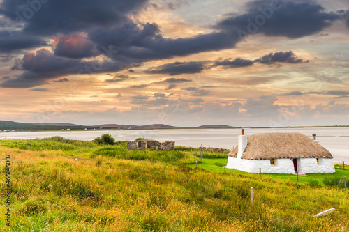 Traditionally built white cottage with thatched roof, next to the turquoise bay, with stormy cloudy dark skies above.island of North Uist, Scotland
