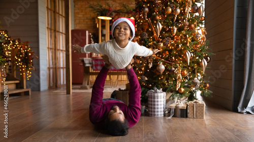Wide banner panoramic view of happy little biracial boy have fun play with loving young father at home. African American dad lying on floor near Christmas tree engaged in funny game with small son.