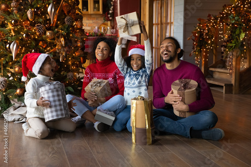 Portrait of happy African American family with two small kids sit on floor near Christmas tree have fun unpacking gifts. Overjoyed biracial parents with little children celebrate New Year together.