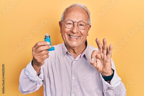 Senior man with grey hair holding medical asthma inhaler doing ok sign with fingers, smiling friendly gesturing excellent symbol