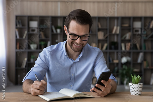 Happy young handsome business man in eyeglasses looking at cellphone screen, web surfing useful information writing notes in paper planner, planning workday or checking working schedule at home office