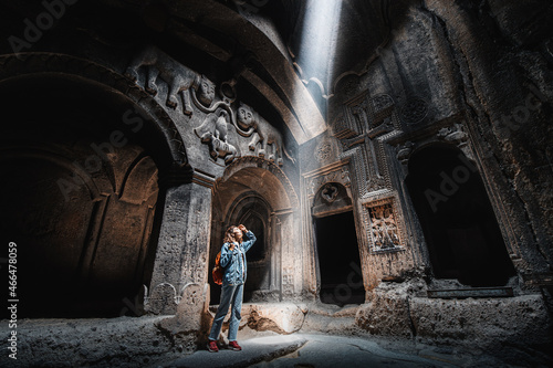 Woman tourist explores the mystical interior of the main hall of the Armenian Geghard Monastery. A ray of light falls on ancient bas-reliefs carved on the walls