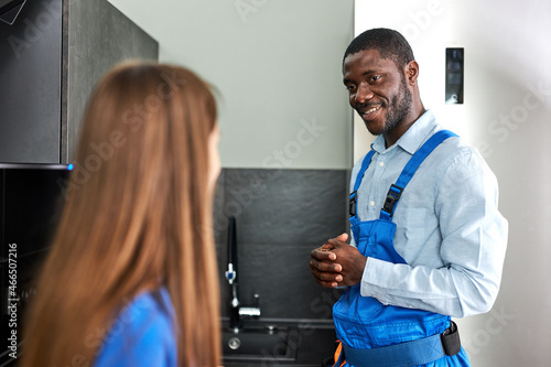 Caucasian Client Woman is grateful to smiling afro plumber for repair glitch in kitchen, confident handyman in blue overalls having talk with female client, discussing repair. focus on plumber