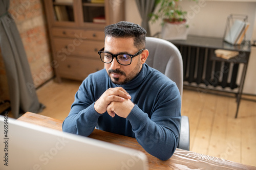 Serious middle-aged teacher or student in eyeglasses sitting in front of computer screen and taking part in online lesson
