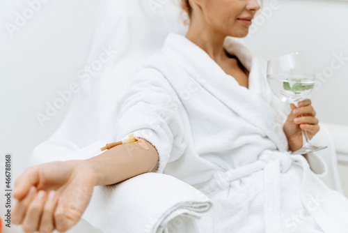 Close up of calm female patient sitting with tube and needle during IV infusion. Girl holding glass with lemon water and relaxing