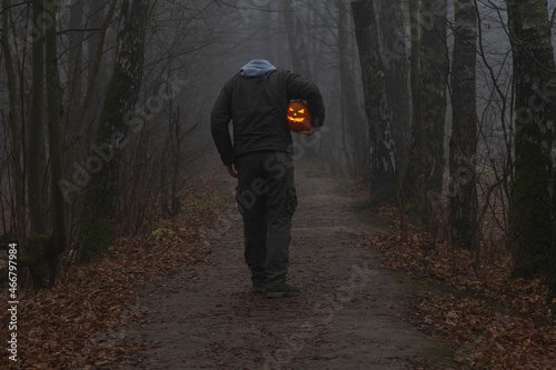 Headless man holds Jack O'Lantern Halloween pumpkin head under his arm and walks on countryside road in autumn foggy forest. Carved face on the pumpkin glows orange. Halloween costume theme.