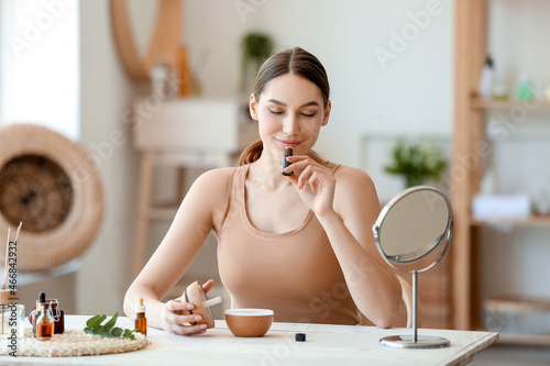 Young woman sitting at table and smelling natural essential oil in bottle