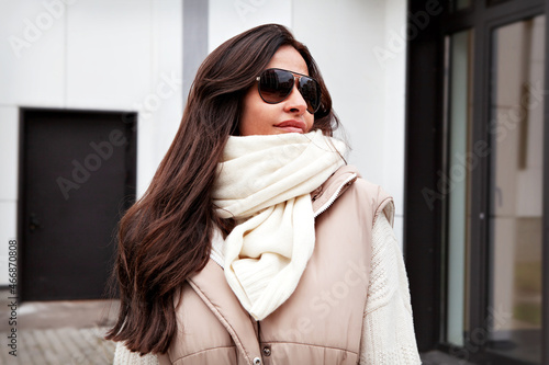 Close up Portrait of beautiful woman looking away on street. Stylish Model with long brunette hair wearing sunglasses, knitted white scarf and sweater, beige vest