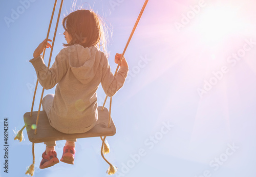 Back view of carefree happy young girl on swing in sunlight against blue sky. Copy space for text or design.