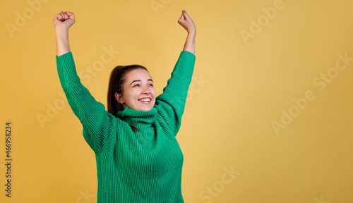 Dynamic image of young cute girl, student wearing green sweater raises hands isolated on yellow studio background. Concept of emotions, fashion