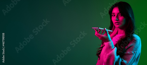Studio shot of young adorable cute girl, student in casual style clothes posing isolated on dark green studio backgroud in pink neon light.