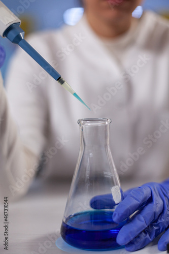 Closeup of specialist researcher dropping medical blue solution in glassware using micropipette working at biological experiment in hospital laboratory. Doctor woman analyzing medicine results