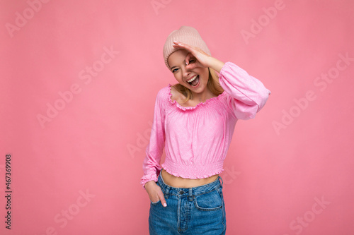 Photo of beautiful happy amusing young blonde female person isolated over pink background wall wearing trendy pink hat and pink blouse looking at camera and showing ok gesture